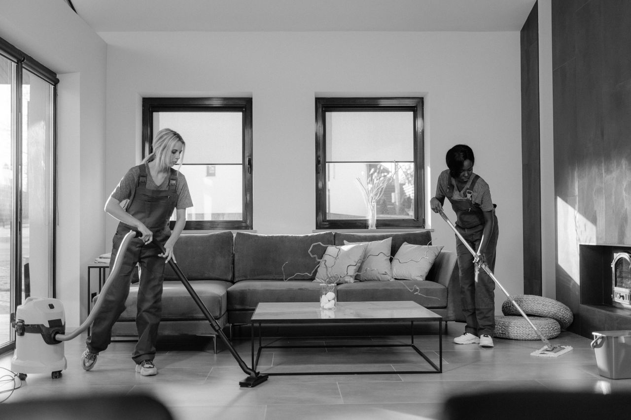 The Art of Drawing Readers In: Your attractive post title goes here Two women in work clothes cleaning a modern living room using a vacuum and mop.